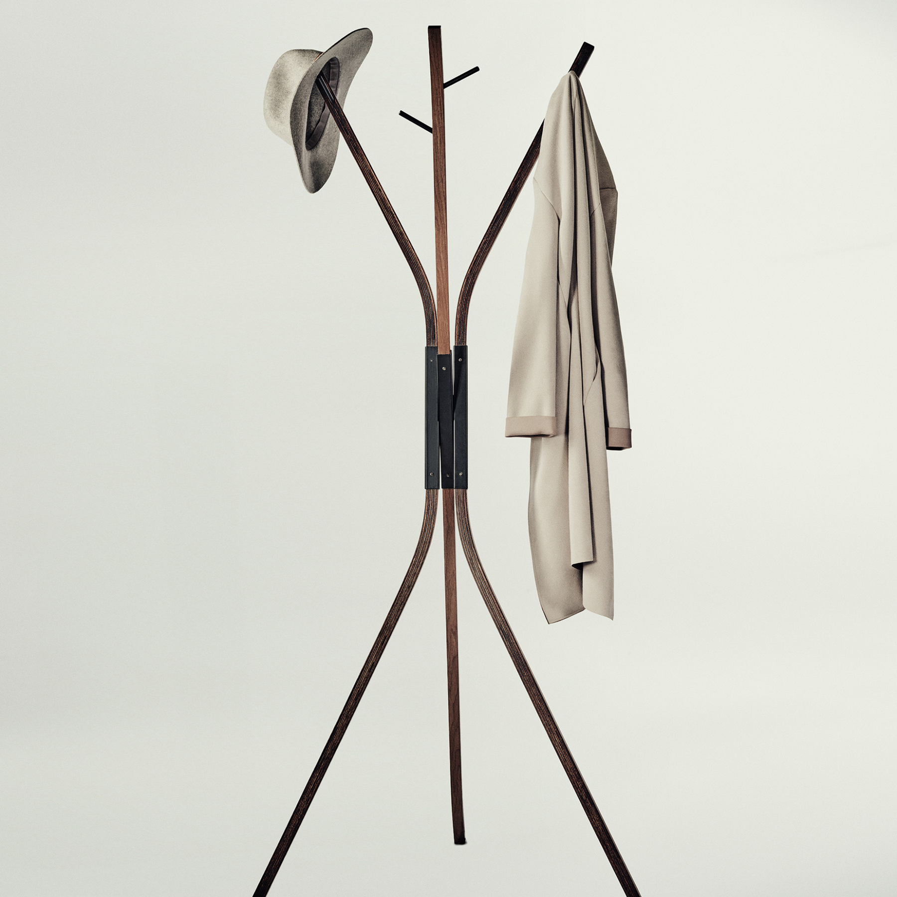A modern wooden coat rack with three legs and curved top arms, shown against a plain light gray background. A hat is hanging on the left hook and a light beige coat is hanging on the right.