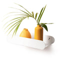 White, capsule-shaped metal shelf with a glossy finish, mounted on a wall with screws. It holds two orange, ribbed vases with green palm leaves, highlighting a minimalist design.