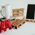 Handmade wooden pallet coasters and phone stands in various colors, including red, natural wood, and dark gray, displayed with a white coffee mug and a smartphone.