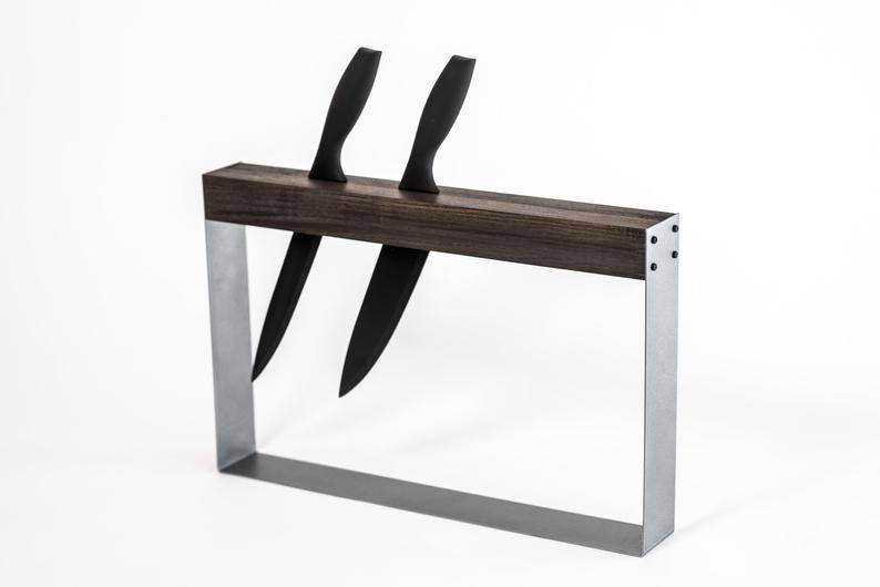 A modern, minimalist knife holder with two black-handled knives inserted vertically into a rectangular wooden and metal frame, designed for easy access and storage.