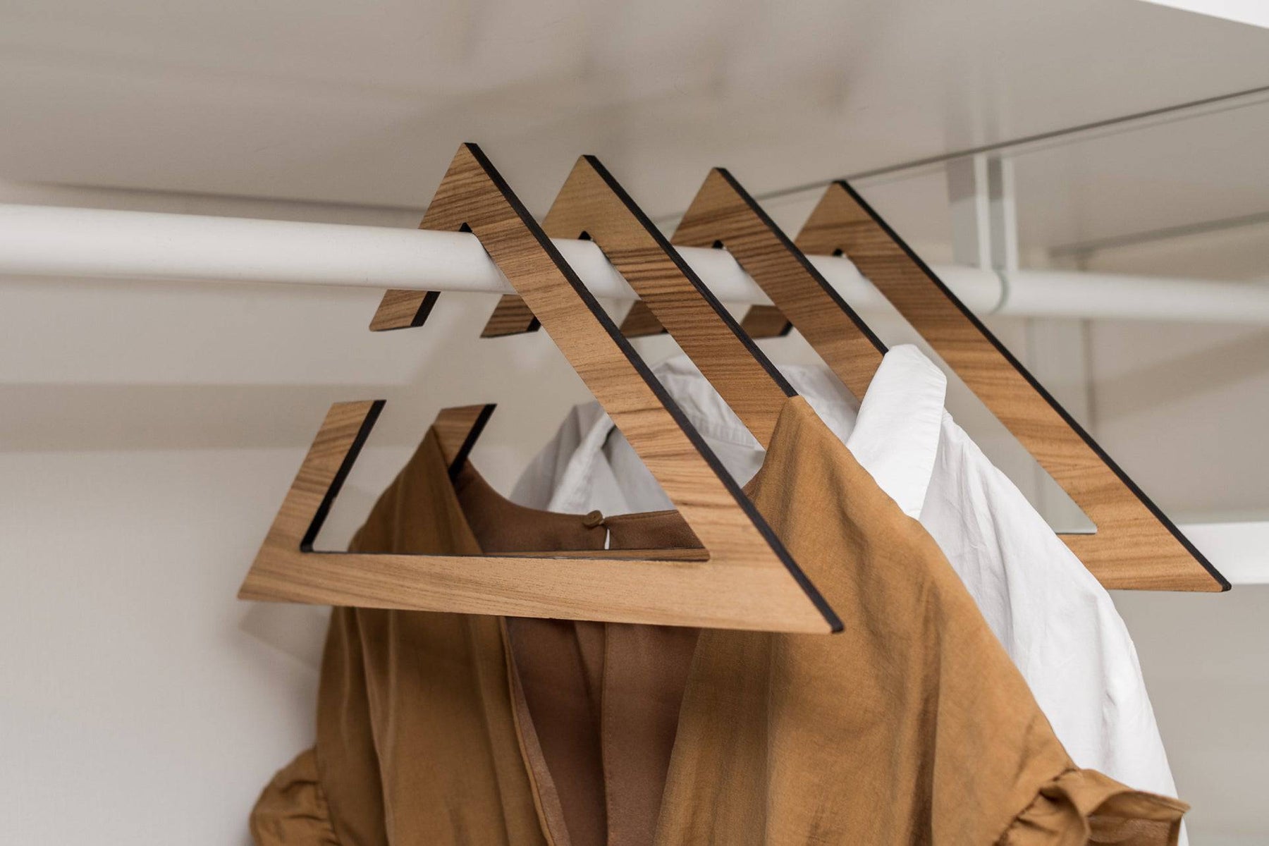 Wooden hangers in paldau finish with a geometric triangular design, holding a brown dress and a white shirt in a closet.