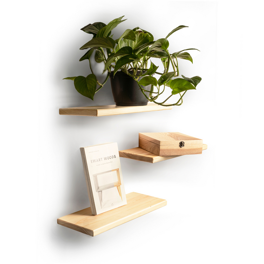 Set of three floating light wood shelves with rounded edges and visible wood grain. The top shelf holds a potted plant, the middle shelf has a wooden box with a latch, and the bottom shelf displays a book titled &