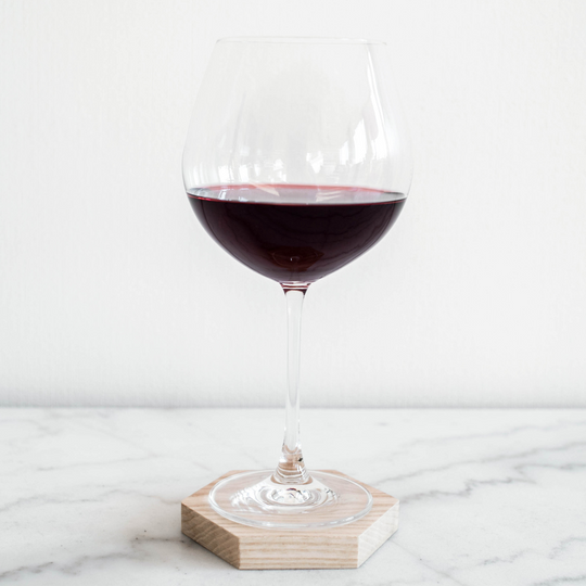 A wine glass filled with red wine, placed on a light-colored hexagonal wooden coaster, set against a minimalist white background on a marble surface.