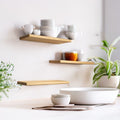 Set of three floating wooden shelves with a natural finish, mounted on a kitchen wall. The shelves hold white ceramic dishes, showcasing their practicality and minimalist design. A plant sits on the countertop below.
