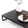 Wooden serving tray with a dark ash finish, featuring a textured surface and sleek metal handles. The tray is styled with a white vase, dried flowers, and a plate with a slice of chocolate cake.