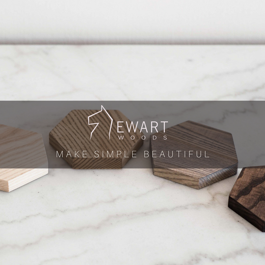 A collection of wooden hexagonal coasters in varying finishes arranged on a light marble surface. The central focus is the Ewart Woods logo, accompanied by the slogan "Make Simple Beautiful,"
