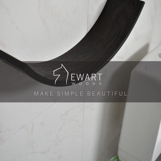 Close-up of a dark wood curved toilet paper holder mounted on a bathroom wall. The EWART WOODS logo and the slogan MAKE SIMPLE BEAUTIFUL are overlaid on the image.