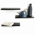 Three floating black wooden ledges with sleek, minimalist design and smooth finishes. The middle ledge holds a modern blue watering can and bottle, while the bottom ledge displays a book titled &