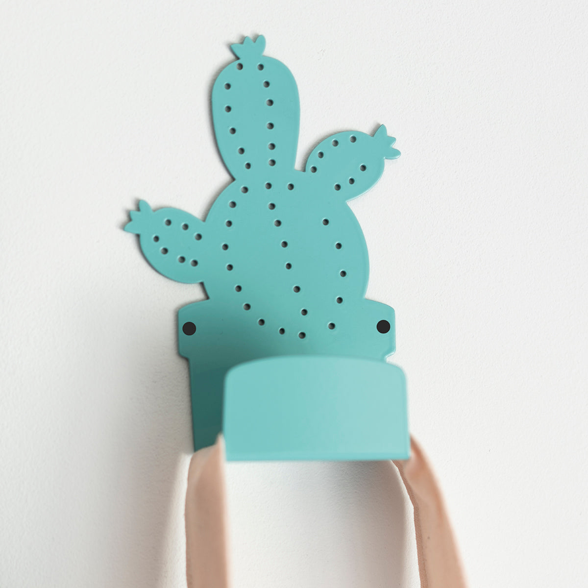 Turquoise metal wall hook in the shape of a cactus, featuring a perforated texture and visible screws, mounted on a white wall, holding a pink tote bag.