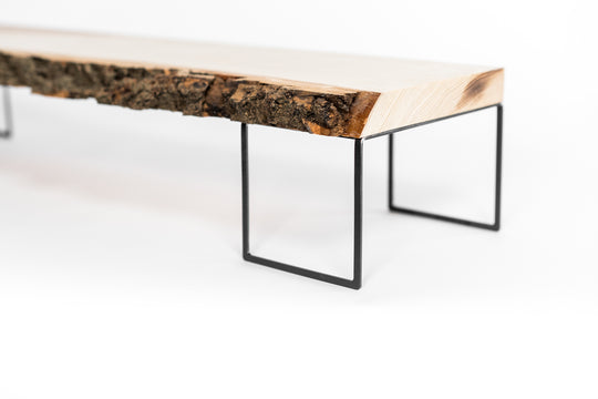 Close-up of a wooden monitor stand with a rough, natural bark edge and a smooth polished top, supported by thin black metal legs.