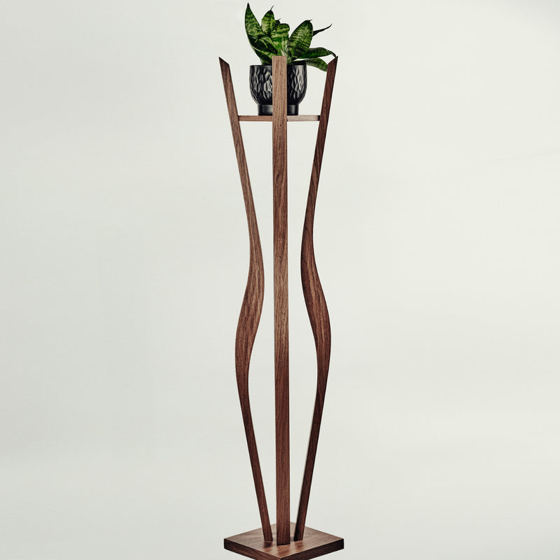 A tall sculptural wooden plant stand with a rectangular base and two curved vertical supports, holding a small potted green plant on a narrow shelf. The stand appears to be made of medium-to-dark brown wood, photographed against a plain light gray studio background.