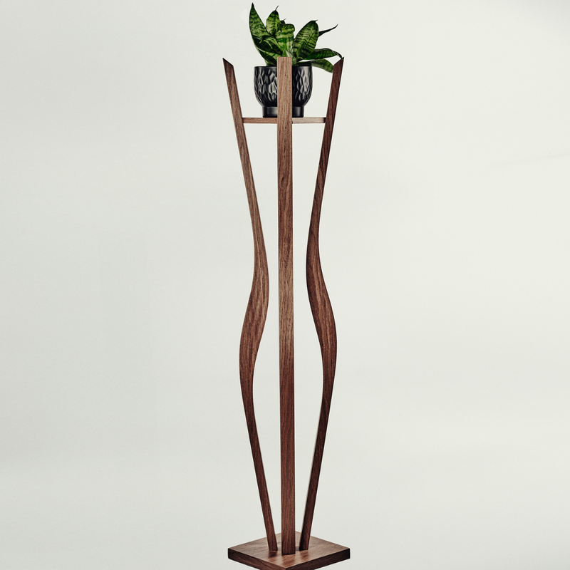 A tall sculptural wooden plant stand with a rectangular base and two curved vertical supports, holding a small potted green plant on a narrow shelf. The stand appears to be made of medium-to-dark brown wood, photographed against a plain light gray studio background.