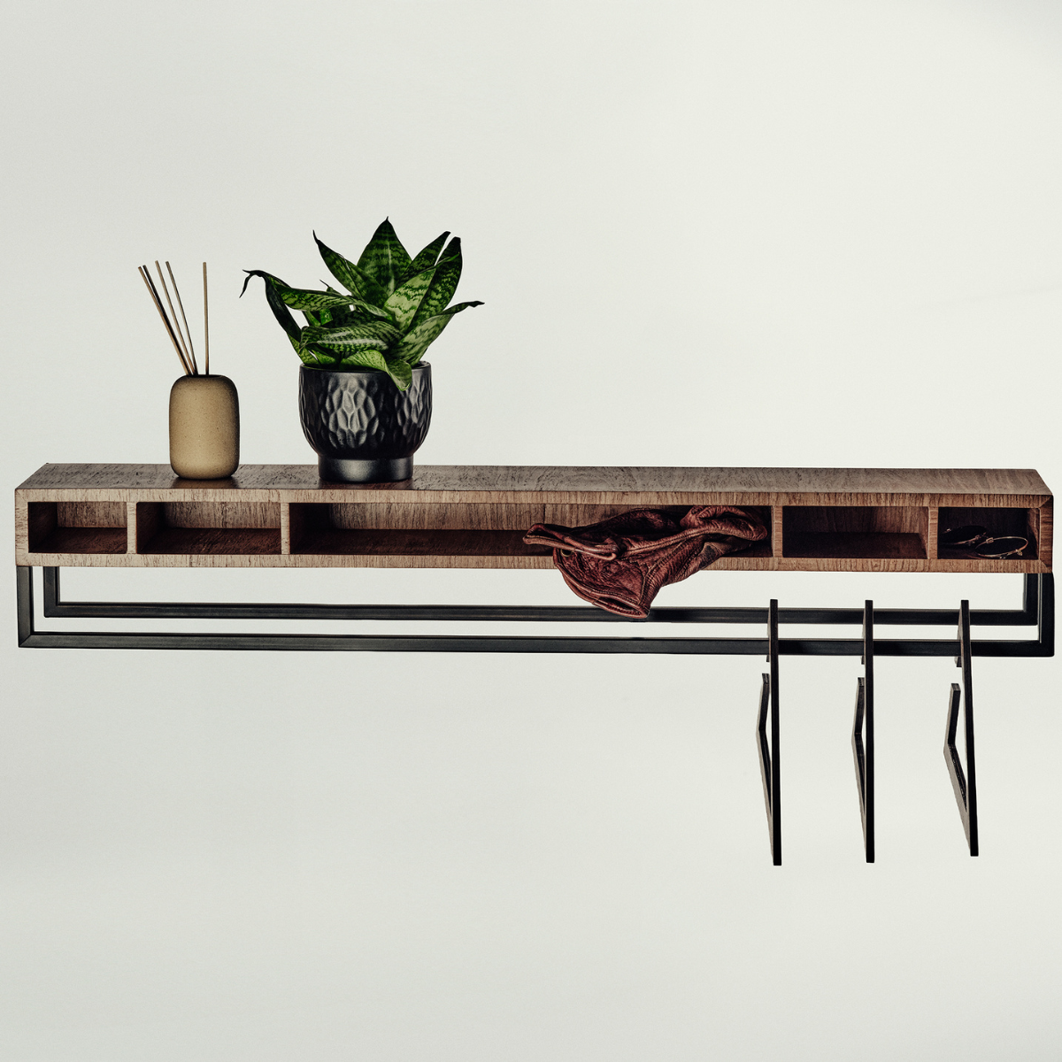 A modern wall-mounted wooden console shelf with open cubbies, a black metal rail, and three hanging hooks, styled with a potted snake plant, a diffuser, and a brown leather accessory against a plain light background.
