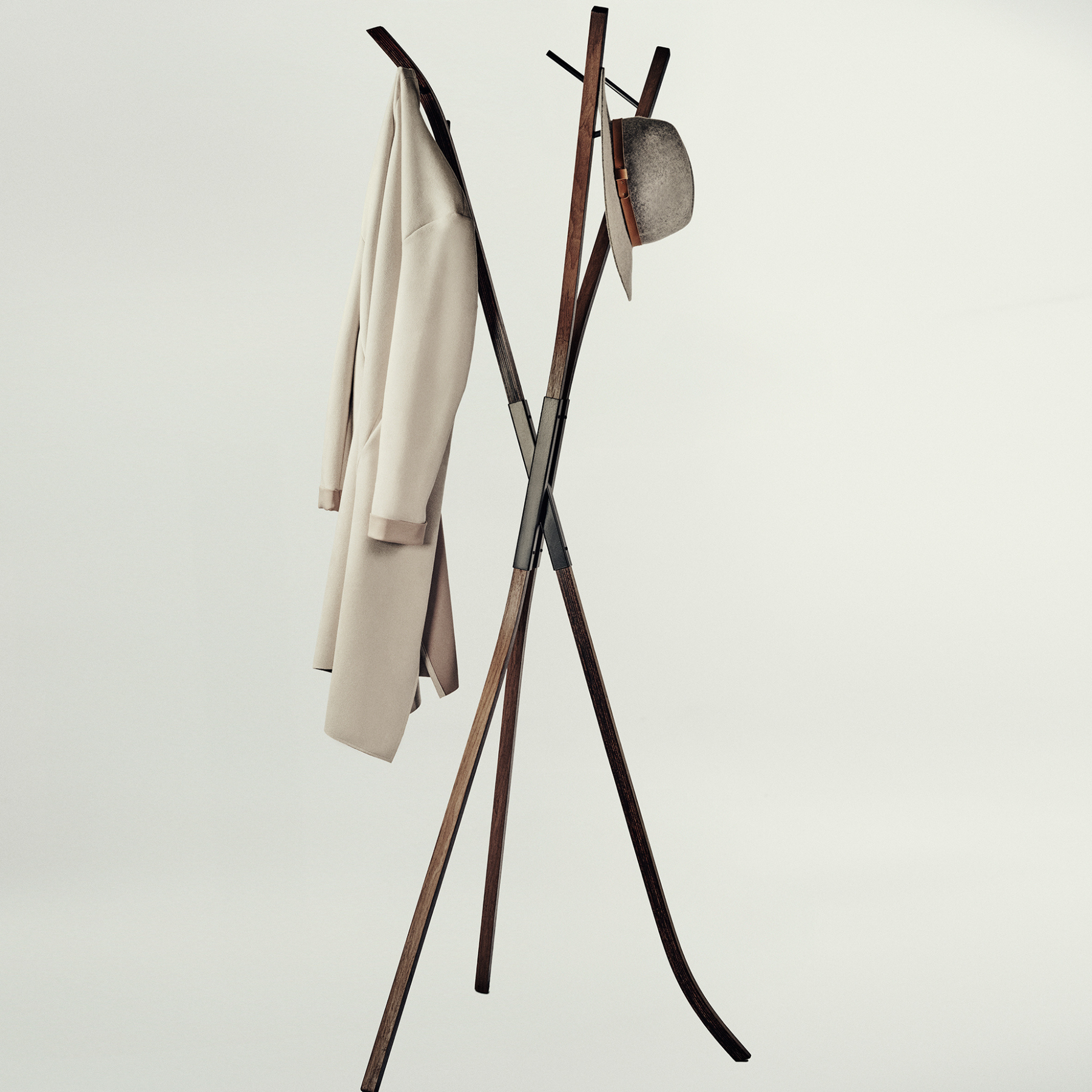 A modern wooden coat rack with an X-shaped base and curved top arms, shown against a plain light gray studio background. A light beige coat hangs on the left hook, and a textured gray hat is suspended on the right.