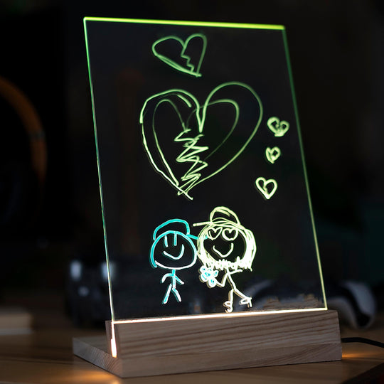 Illuminated acrylic panel in a wooden base with neon-colored drawings of two smiling figures, a broken heart, and smaller hearts, glowing against a dark background.