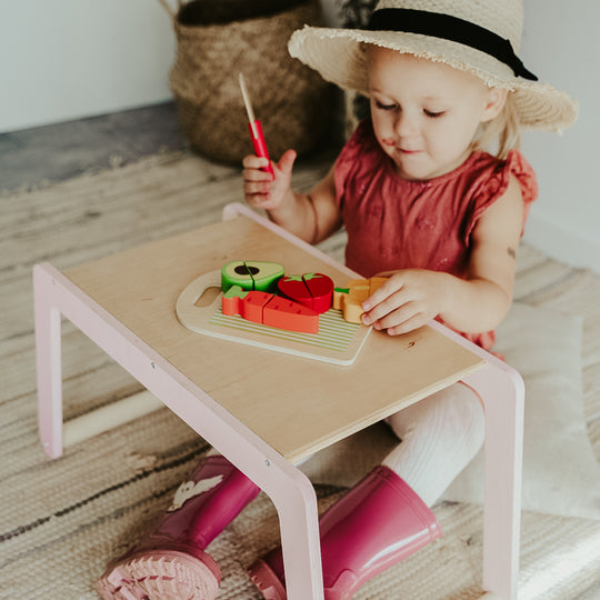Young child in a sunhat and coral dress plays with wooden toy food at a pastel pink and natural wood activity table, wearing bright pink rain boots.