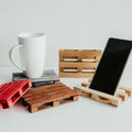 A collection of small wooden pallet coasters in various colors, holding a white mug and a smartphone. The coasters come in red, gray, natural wood, and other stained finishes, displayed on a white surface.