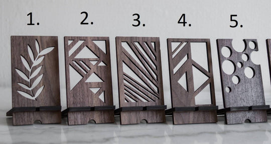 Wooden phone stands in walnut finish with unique geometric cutout designs: 1. Leaf pattern, 2. Abstract triangles, 3. Diagonal lines, 4. Geometric shapes, 5. Circular cutouts. Each stand holds a phone at an angle on a smooth surface.
