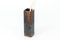 Tall, rectangular utensil holder with a rustic copper finish and wooden base, holding a pair of wooden utensils. The holder is displayed against a plain white background.