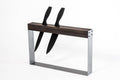 A modern, minimalist knife holder with two black-handled knives inserted vertically into a rectangular wooden and metal frame, designed for easy access and storage.