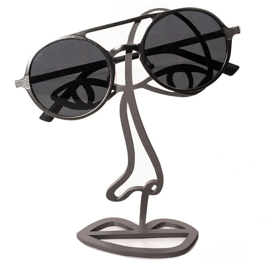 Black metal abstract face sculpture for glasses. The minimalist design features a wireframe outline of eyes, nose, and lips, with a pair of black sunglasses being held on the upper part.