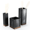 Set of three black metal rectangular utensil holders with wooden bases in varying heights, each holding light-colored wooden utensils, against a plain white background.