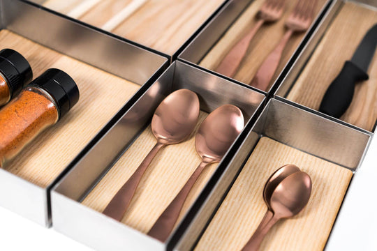 Organized metal trays containing various kitchen utensils, including two copper spoons, forks, a knife, and spice containers filled with red seasoning. The utensils are neatly arranged on a wooden surface.