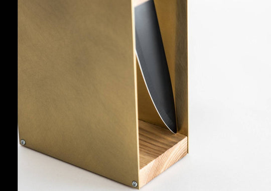 Close-up of a gold knife block with a natural wood base and a visible side slot holding a knife. The smooth wooden grain contrasts with the matte green metal, displayed against a white background.