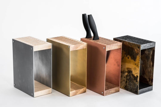 Four metal knife blocks in different finishes: silver/aluminum, gold/brass, rosegold/copper, and vintage black/rustic steel. Each block has a wooden top and base, displayed side by side against a plain background.