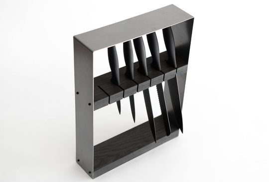 Angled view of a minimalist black knife block with a dark wooden top and metal frame, holding six black-handled knives in horizontal slots, displayed against a white background.
