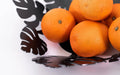 Close-up of a sleek black metal bowl featuring tropical leaf cutouts, filled with fresh, vibrant oranges.