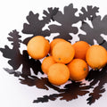 Black metal decorative bowl with leaf-shaped cutouts, holding several bright orange fruits.