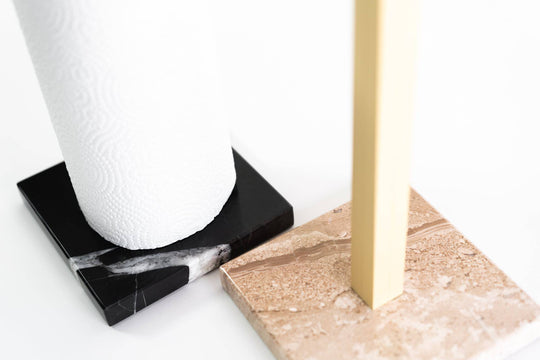 Close-up of marble-based paper towel holders, one with a black marble base and white roll, the other with a beige marble base and gold metal stand, showcasing their sleek and luxurious design.