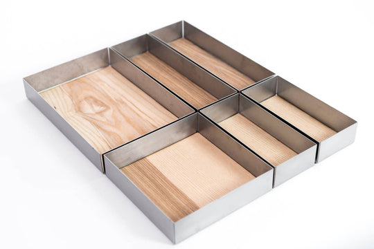 Empty modular kitchen drawer organizers made of metal and wood, consisting of six rectangular compartments of various sizes. The dividers rest on a light wooden base, designed for organizing utensils or kitchen items.