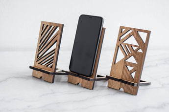 Wooden phone stand set in walnut finish with a geometric pattern, holding a phone in the center, placed on a smooth surface. The stands are designed with modern, angular shapes on each side.