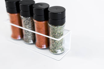 Spice rack organizer with white metal frame, holding four spice jars with black caps, neatly arranged in a row.
