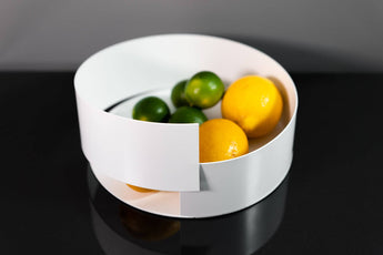 Modern white metal bowl with a sleek, overlapping design, holding vibrant lemons and limes, displayed on a reflective black surface.
