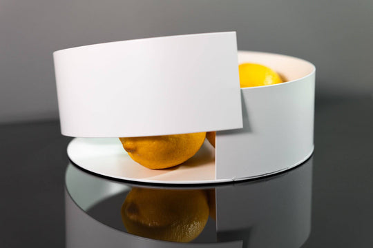 Modern white metal bowl with a sleek, overlapping design, holding vibrant lemons and limes, displayed on a reflective black surface.