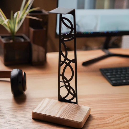 Modern headphone stand with a sturdy birch wood base and sleek black metal frame featuring artistic cutout patterns, placed on a wooden desk in a minimalistic office setup.
