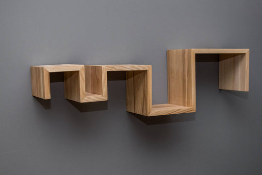 Zigzag-shaped floating shelf made of light-colored wood, mounted on a gray wall. The shelf features clean, sharp edges and an angular design with multiple levels.