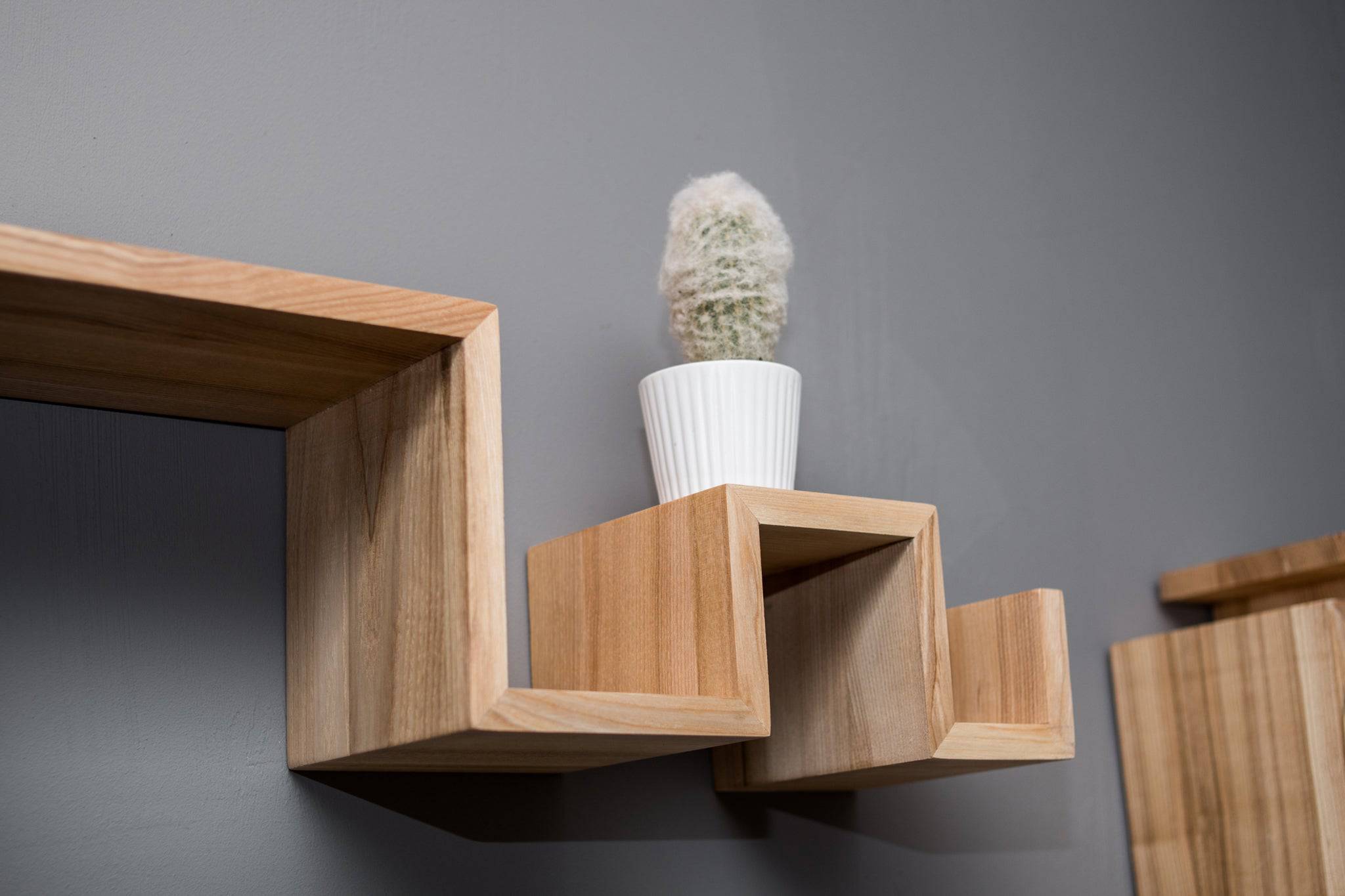 Close-up of a zigzag shelf made of light-colored wood, with clean, sharp edges, mounted on a gray wall. The shelf holds a small potted cactus.