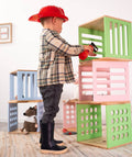 A child wearing a firefighter helmet and boots holds a toy fire extinguisher aimed at a stack of colorful wooden blocks with various cut-out designs, including slats and circular holes.