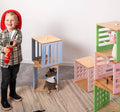 Child dressed as a firefighter holding a toy fire extinguisher, surrounded by colorful stacked wooden blocks in blue, pink, and green, with cut-out grids and circles. Stuffed animals are placed inside the blocks.