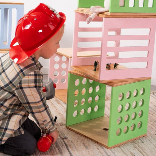 A child wearing a red firefighter helmet plays with a set of colorful wooden building blocks designed to resemble structures, complete with toy soldiers inside.