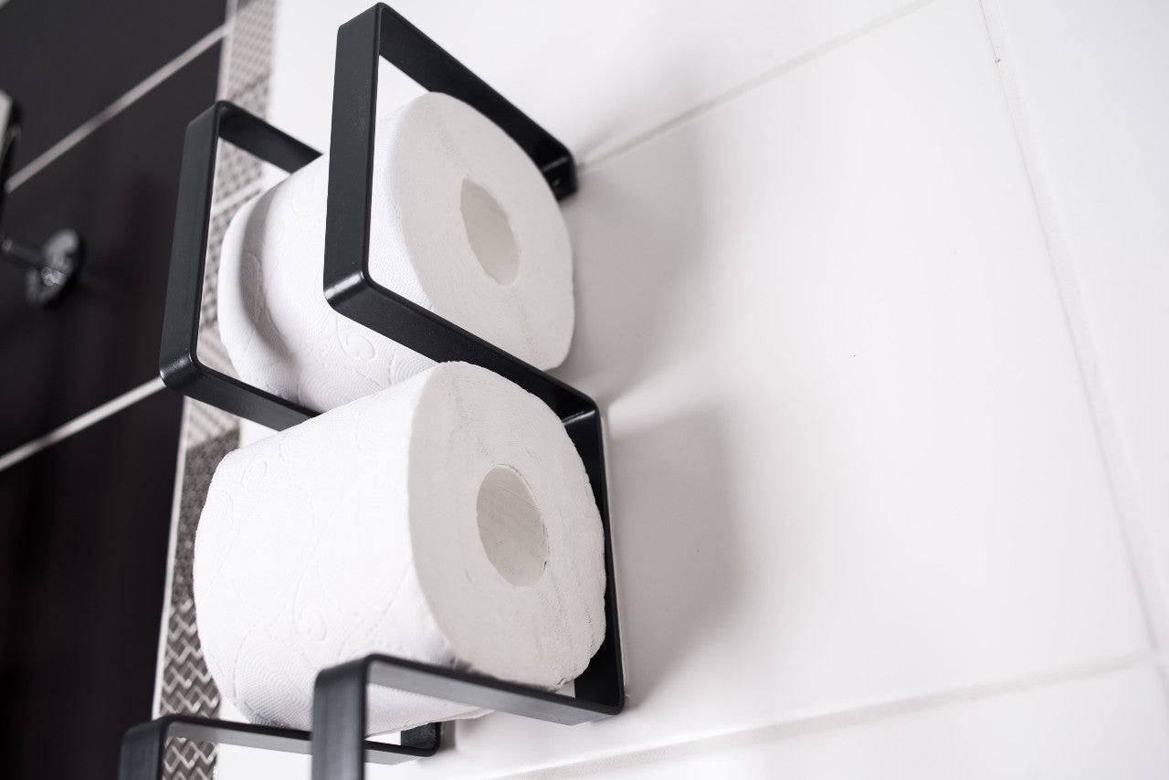 Close-up of a wall-mounted black toilet paper holder with a geometric frame, holding two rolls of toilet paper in a vertical arrangement.