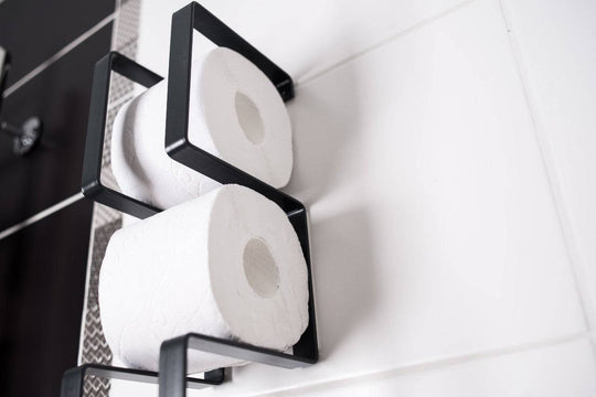 Close-up of a wall-mounted black toilet paper holder with a geometric frame, holding two rolls of toilet paper in a vertical arrangement.