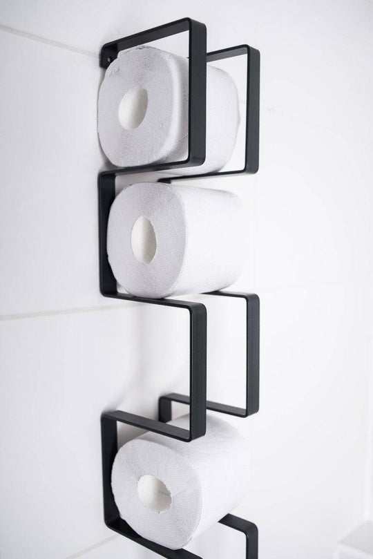 Close-up of a wall-mounted black toilet paper holder with a geometric design, displaying three rolls of toilet paper stacked vertically.