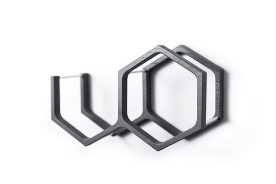 Close-up of a wall-mounted black mini honeycomb toilet paper holder. The holder features metal bars for support and a geometric design with textured wood.